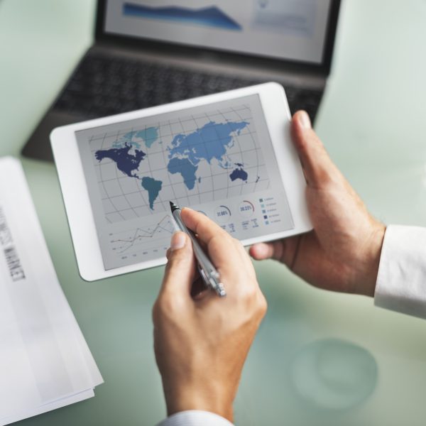 Plano detalhado de mãos a segurar um tablet que exibe um mapa-múndi e gráficos de análise de mercado global