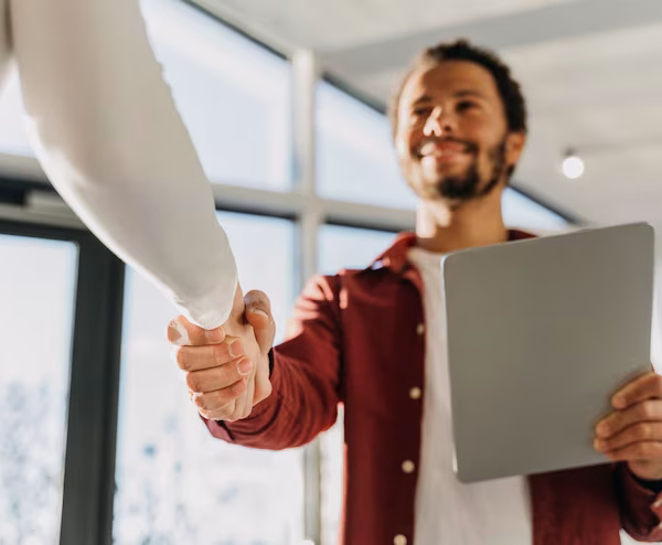 Plano de ângulo baixo de um homem jovem e sorridente a segurar um tablet e a cumprimentar alguém com um aperto de mão num ambiente de trabalho luminoso.