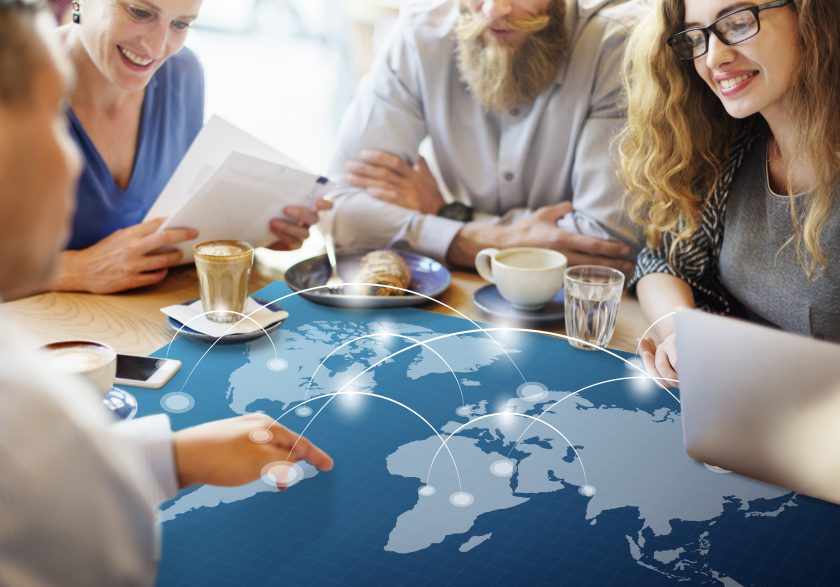 Grupo de profissionais numa reunião informal à volta de uma mesa, com uma sobreposição gráfica de um mapa-múndi e linhas de conexão digital