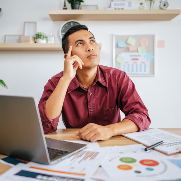 Homem de camisa bordeaux sentado à secretária com um computador portátil, a olhar para cima pensativo enquanto analisa documentos e gráficos espalhados na mesa.