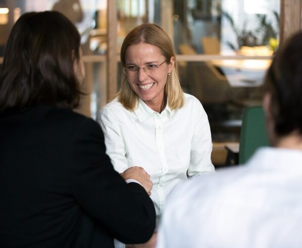 Mulher de negócios sorridente com óculos e camisa branca a cumprimentar um colega com um aperto de mão durante uma reunião de trabalho num escritório moderno.
