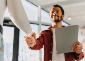 Plano de ângulo baixo de um homem jovem e sorridente a segurar um tablet e a cumprimentar alguém com um aperto de mão num ambiente de trabalho luminoso.