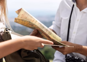 Plano aproximado de um casal a consultar um mapa de papel ao ar livre, com uma mochila e uma câmara fotográfica pendurada ao pescoço.