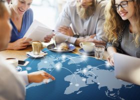 Grupo de profissionais numa reunião informal à volta de uma mesa, com uma sobreposição gráfica de um mapa-múndi e linhas de conexão digital