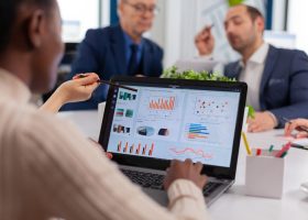 Equipa multidisciplinar numa reunião de trabalho sobre energias limpas, com maquetes de turbinas eólicas, painel solar fotovoltaico e análise de mapas topográficos.